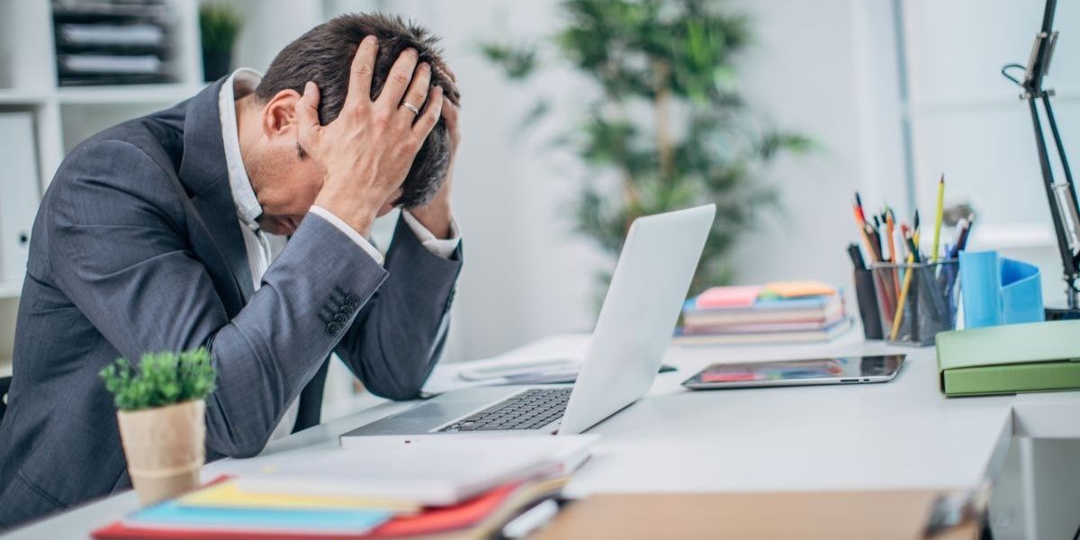 Frustrated businessman sitting at his desk with his head in his hands, symbolizing the stress and financial strain caused by non-producing managers affecting mortgage broker compensation.