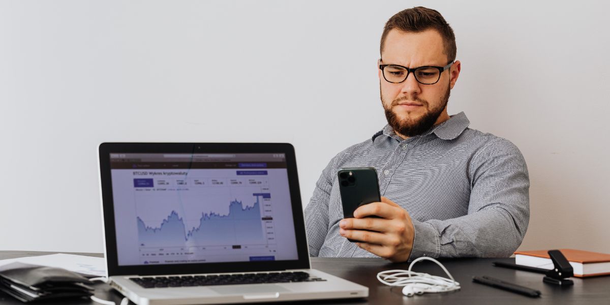 A man using a phone and sitting in front of a laptop with a graph
